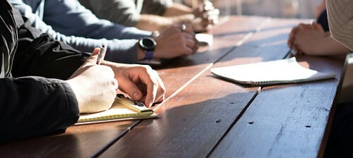 A group of team members meeting around a wooden table, taking notes and discussing a project during a collaborative planning session.